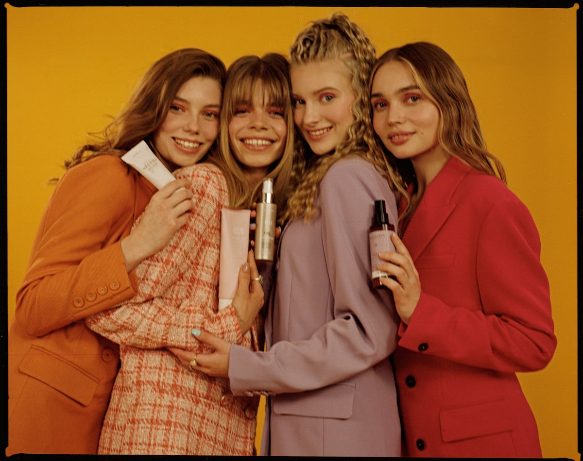 Four smiling women holding beauty products against a yellow background.